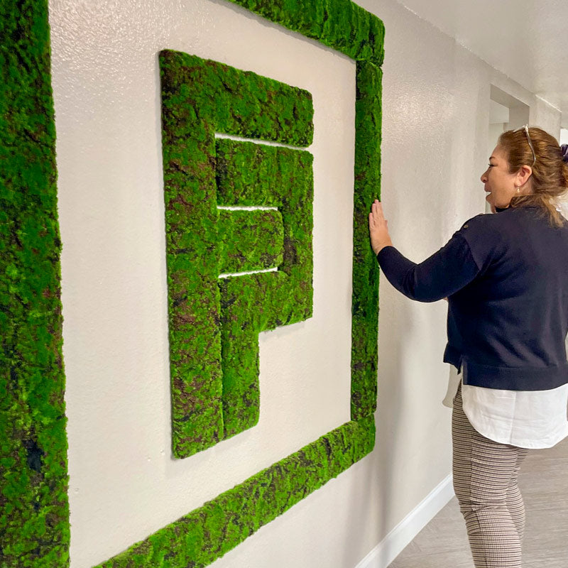 Woman in blue sweatshirt touching moss green wall logo on a white wall.