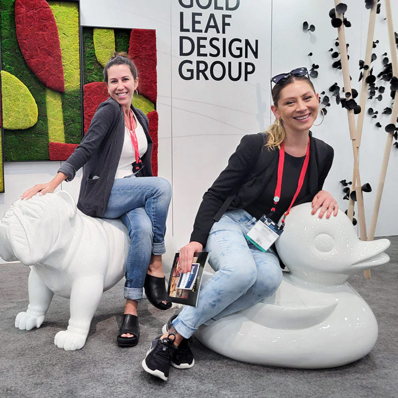 Two people sitting large white fiberglass sculptures (a bulldog and a duck) at the Gold Leaf Design Group HDExpo booth.